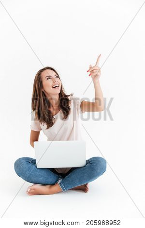 Vertical image of happy brunette woman holding laptop computer and pointing up while sitting on the floor over white background
