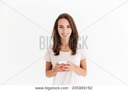 Smiling brunette woman holding smartphone and looking at the camera over white background