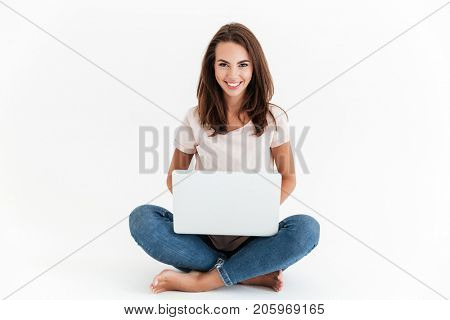 Smiling brunette woman holding laptop computer and looking at the camera while sitting on the floor over white background