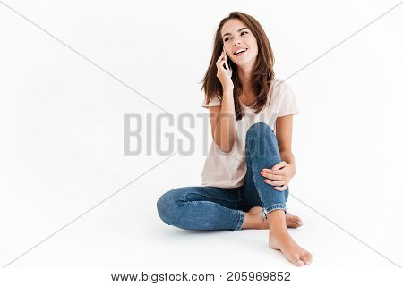Smiling brunette woman sitting on the floor and talking by the smartphone over white background