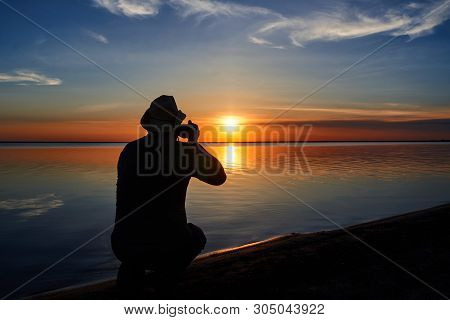 A Man Photographs The Sunset Over The Water And The Sky With Clouds.