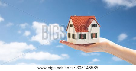 real estate, accommodation and property concept - close up of hand holding house or home model over blue sky and clouds background