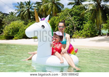 Smiling Portrait Of Mother And Daughter Enjoying Sitting On White Unicorn Float At Beach