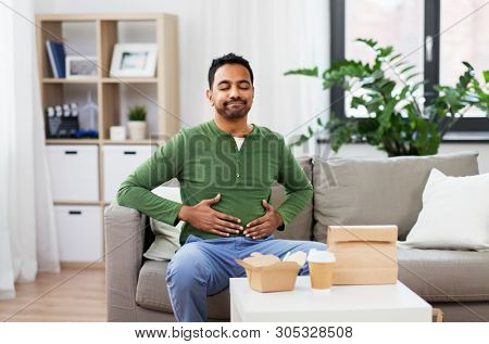 consumption and people concept - full and pleased indian man eating takeaway food at home