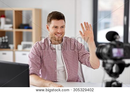 blogging, videoblog and people concept - male blogger with camera recording video and waving hand at home office
