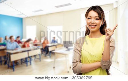 school, education and learning concept - happy asian female teacher or student girl pointing finger up over classroom background