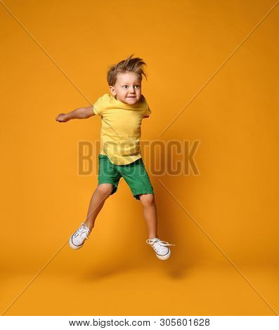 Full Length Portrait Of An Excited Young Boy Jumping Leaping In Green Shorts Over Yellow Background