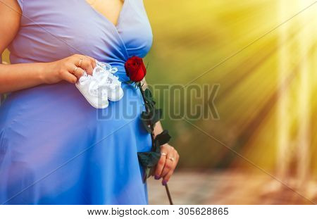 Pregnant Woman Holding White Baby's Bootee Wearing In Blue Dress Over Nature Park Background At Suns