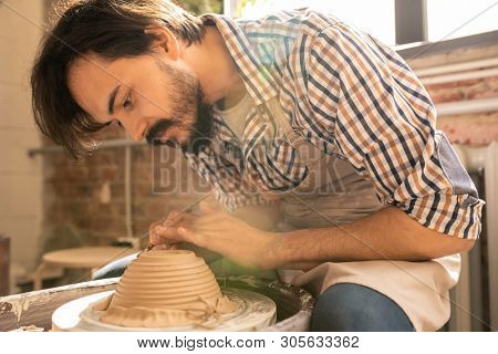 Young creative artisan bending over rotating clay item on wheel while using one of handtools for cutting and ornamenting