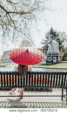 A Girl In A Floral Dress Sitting On A Park Bench Holding A Red Umbrella Surrounded By Red Tulips Wit