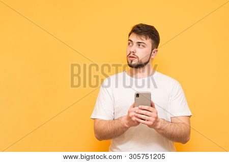 Calm Man With A Beard On A Yellow Background, Uses A Smartphone, Looking Sideways To The Empty Space
