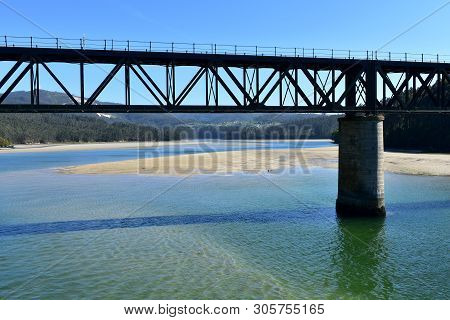 Beach With Clear Water, Forest And Train Bridge. O Barqueiro, Spain.