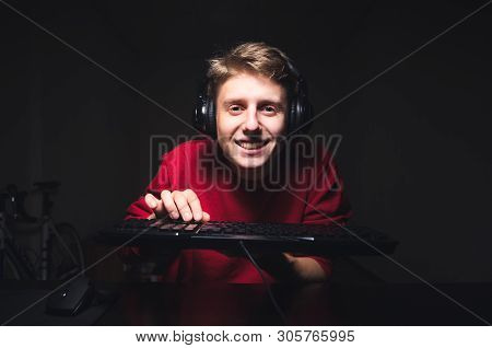 Smiling Gamer Plays Video Games Against The Backdrop Of A Dark Room, Holds The Keyboard In His Hand,