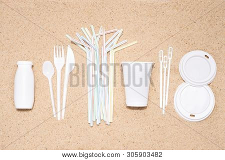 Disposable single use plastic objects such as bottles, cups, forks, spoons and drinking straws that cause pollution of the environment, especially oceans. Top view on sand
