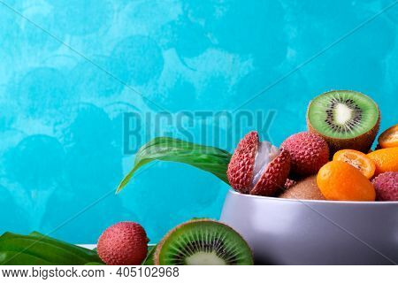 Lychee, Kiwi And Kumquat In A Gray Ceramic Bowl Against The Turquoise Background. Assorted Tropical 