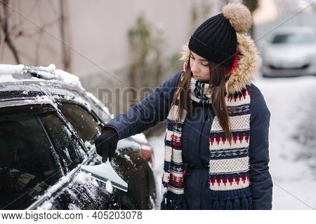 A Woman Is Cleaning Snowy Window On A Car With Snow Scraper. Pretty Woman Warmy Dressed Clean Her Ca