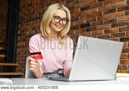 Glad Mid Age Woman Using A Laptop For E-bancing, Inputs Credit Card Number To Make Online Transactio