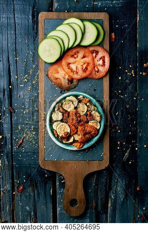 high angle view of a rustic green plate with some slices of dried tomato and zucchini, served as appetizer or snack, and some slices of raw tomato and zucchini, on a dark gray rustic wooden surface