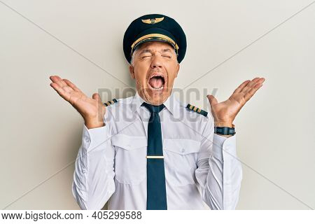 Handsome middle age mature man wearing airplane pilot uniform celebrating mad and crazy for success with arms raised and closed eyes screaming excited. winner concept 