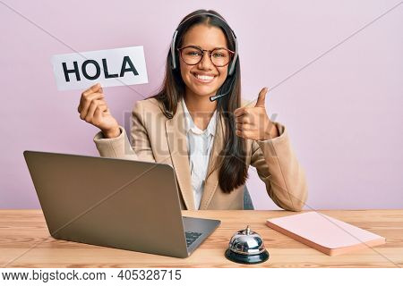 Beautiful hispanic woman wearing operator headset holding hola greenting smiling happy and positive, thumb up doing excellent and approval sign 