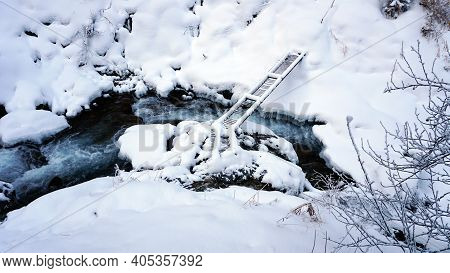 A Clear Mountain River Runs Through A Snowy Gorge. There Is A Metal Bridge Covered With Snow. Tall S