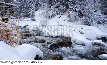 A Clear Mountain River Runs Through A Snowy Gorge. There Is A Metal Bridge Covered With Snow. Tall S