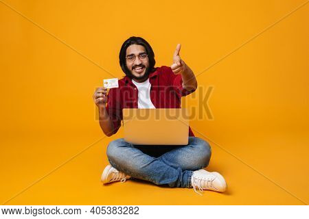 Happy indian man using laptop for shopping online with credit card while sitting isolated over yellow background, humbs up