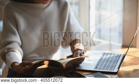 Closeupl Woman Reading The Book In Hands While Sitting At The Wooden Working Desk With Computer Lapt