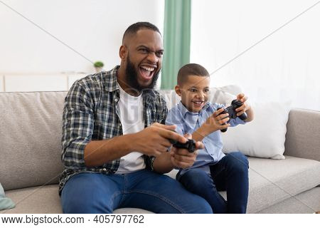 Excited African Dad And Son Playing Game Having Fun Sitting On Couch At Home. Young Black Father And