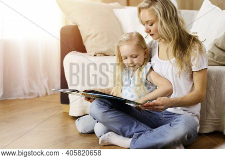 Happy Family. Blonde Young Mother Reading A Book To Her Cute Daughter While Sitting At Wooden Floor 