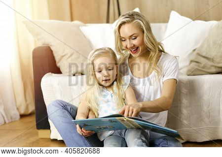 Happy Family. Blonde Young Mother Reading A Book To Her Cute Daughter While Sitting At Wooden Floor 