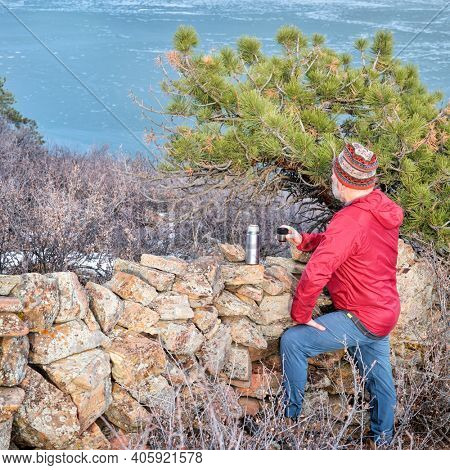 senior hiker is taking a tea break at old stone fence overlooking frozen mountain lake  - Horsetooth Reservoir at foothills of Rocky Mountains - a popular recreational area in northern Colorado