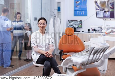Healthcare Specialist In Teeth Hygine Wearing White Coat Sitting On Stool And Assistant Discussing W