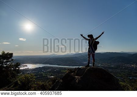 Silhouette Of Girl In Hat, Stains On A Rock And Spreads Her Hands To The Sides. A City, Mountains, S
