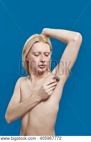 Young Caucasian Man With Long Fair Hair Shaving Armpit Using Razor While Standing On Blue Background