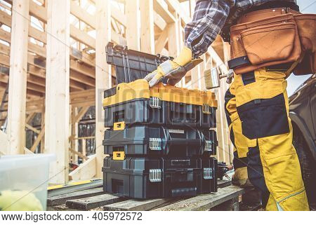 Construction Worker Preparing His Large Plastic Heavy Duty Toolboxes. Wooden Skeleton Frame Of House
