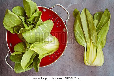 Three Pok Choi Cabbage In Red Colander On Gray Background, Top View