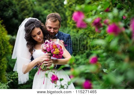 Happy Groom Hugging Beautiful Bride With Bouquet From Behind Near Flower Bush