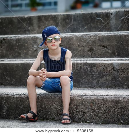 Portrait Of Fashionable Little Boy In Sunglasses And Cap Siting