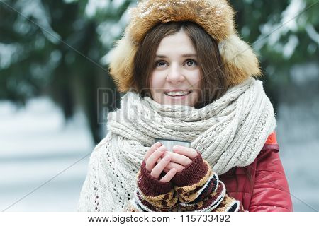 Outdoor winter portrait of cheerful young lady holding tourist vacuum flask cup with hot drink