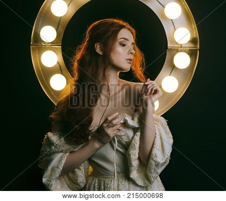 Portrait of a young red-haired woman in a vintage ash dress with open shoulders against a background of lights. A princess. Fairy tale. Art photo