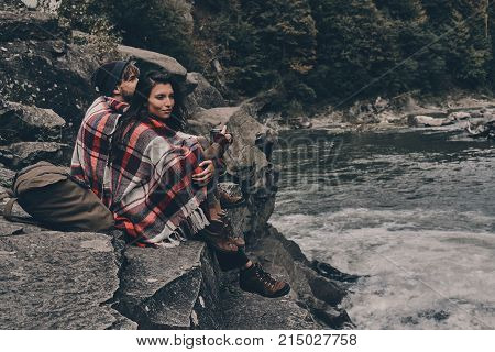 Listening to the song of nature. Beautiful young couple covered with blanket looking away while sitting on the rocks near the river