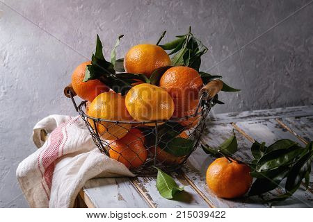Ripe organic clementines or tangerines with leaves in basket standing with kitchen towel on white wooden plank table with gray wall as background. Rustic style. Healthy eating