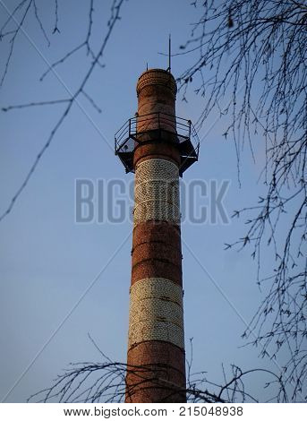 The old chimney against the blue sky in the foreground of the birch branch. The pipe material is red brick. The color of the pipe is red-white. On the top of the pipe there are signal lights.