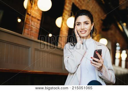 Graceful female with dark hair and eyes sitting in cafe using free internet conncetion while listening to music with earphones and mobile phone. Elegant lady using modern technologies indoors