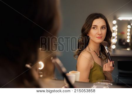 Attractive Girl Primp With Make Up Brush At The Mirror With Lamps In The Beauty Studio.