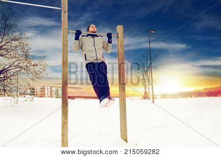 fitness, sport, exercising, training and people concept - young man doing pull ups on horizontal bar outdoors in winter