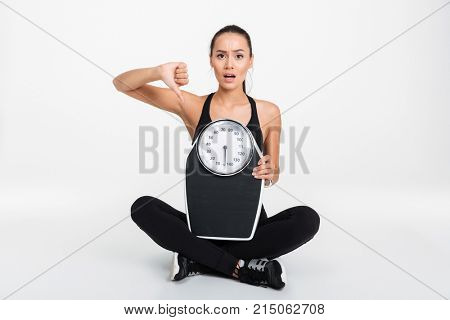 Portrait of a disappointed asian fitness woman holding weight scales and showing thumbs down gesture isolated over white background