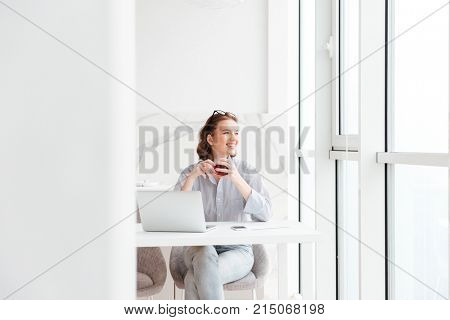 Young attractive brunette woman holding cup of tea while sitting at kitchen and looking at large window