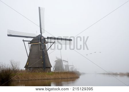 Silhouette Of A Dutch Windmill Under A Misty And Foggy Weather A Long A Calm And Quiet Canal